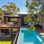 Modern luxury home with pool, timber deck, outdoor dining area, and glass walls surrounded by trees.