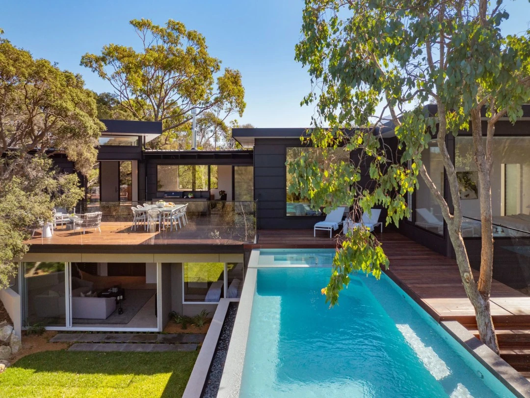 Modern luxury home with pool, timber deck, outdoor dining area, and glass walls surrounded by trees.
